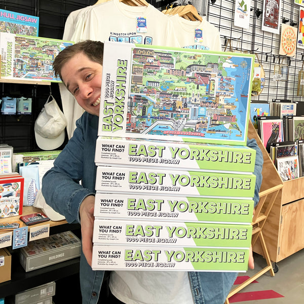Person holding a stack of 'East Yorkshire' jigsaw puzzle boxes in a store setting.