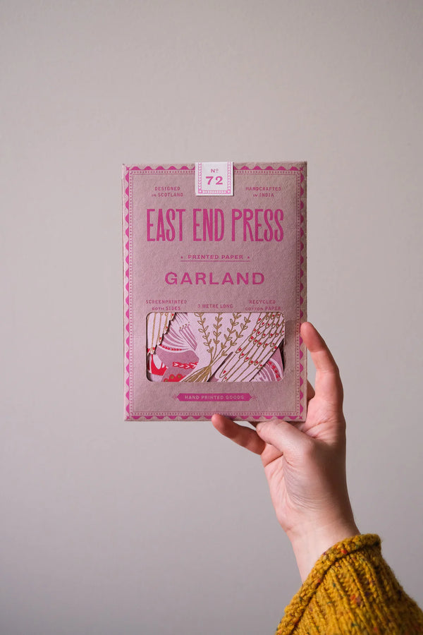 Hand holding a pink box labeled "East End Press Printed Paper Garland," featuring illustrations and text about screen printing and recycled materials.